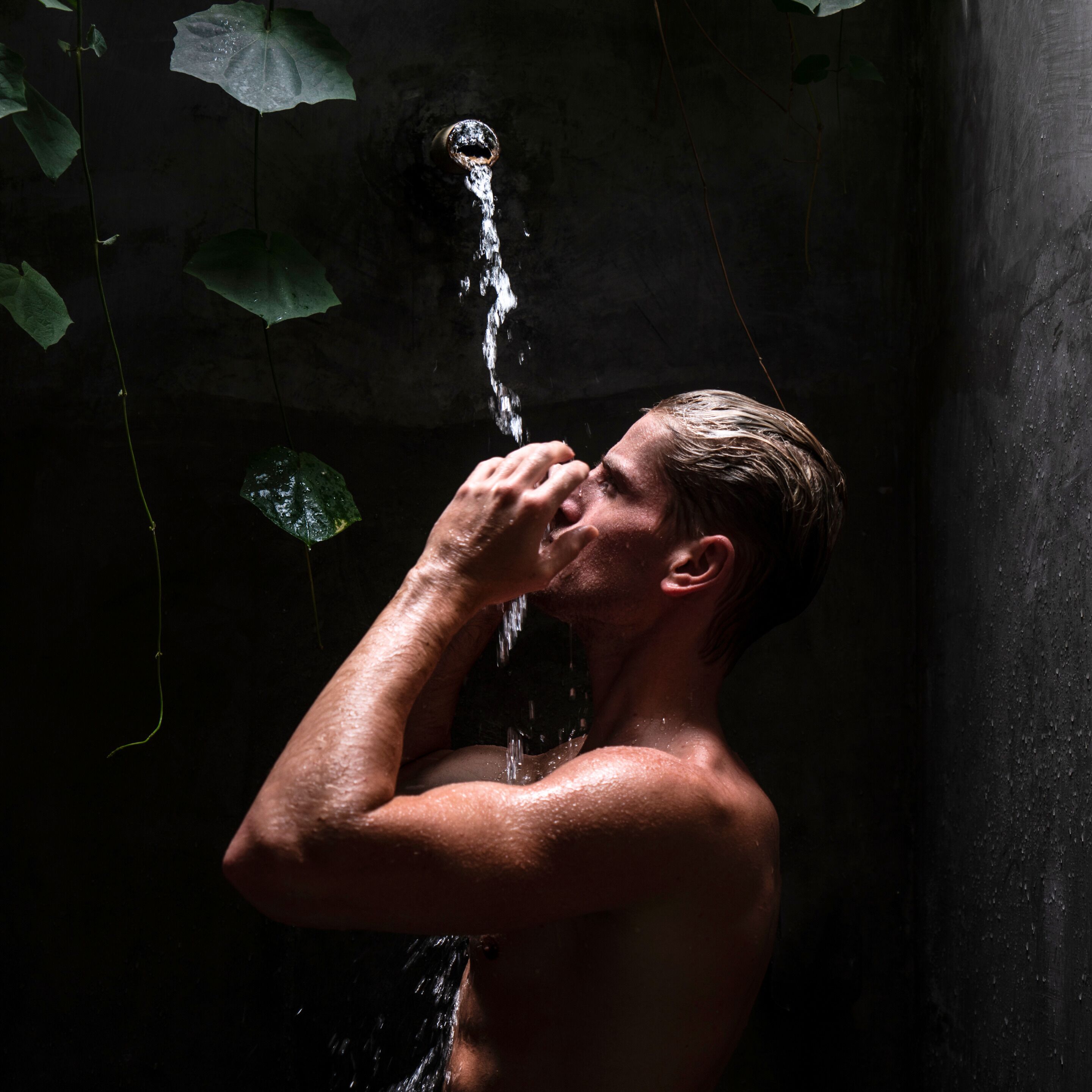 A person drinking water from a stream in a dark, enclosed space with visible foliage.