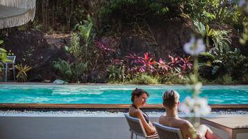 A poolside area with two lounge chairs, a wooden deck, and a natural backdrop of rocks and vegetation.
