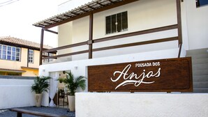 Un letrero de madera con la inscripción "POUSADA DOS Anjos", ubicado frente a un edificio de fachada blanca y techo marrón.