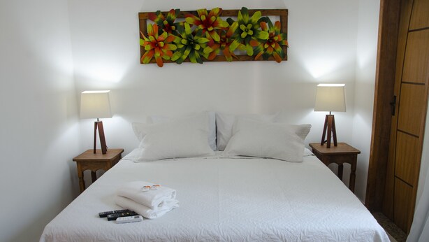 Una cama bien hecha con sábanas blancas, dos lámparas de noche, un cabecero de madera y una obra de arte floral sobre la cama.