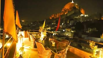 Terraza en la azotea con mobiliario de mimbre y plantas en macetas, con vistas a un paisaje urbano nocturno.