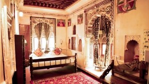 A traditional room with a bed, a red patterned rug, ornate curtains, and a mirror with a decorative frame. - Singhvi's Haveli (Jodhpur)