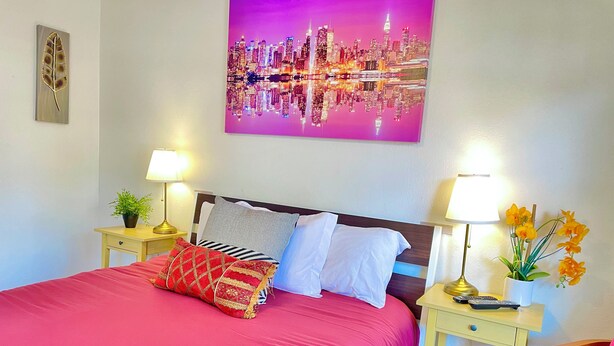 A neatly made bed with a red bedspread, two bedside tables with lamps, a cityscape painting on the wall, and a small potted plant on the nightstand. - New Studio  - Minutes to Convention Center, Disney,Universal ,INTL DR (Orlando)