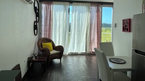 Ein Zimmer mit Esstisch, einem Stuhl, einer Couch mit gelbem Kissen und Blick ins Grüne durch das Fenster.