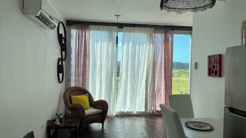 Una habitación con una mesa de comedor, una silla, un sofá con un cojín amarillo y vistas a zonas verdes a través de la ventana.