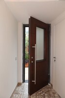 Un pasillo con puerta de madera, suelo de baldosas y vista a un jardín a través de una puerta abierta.