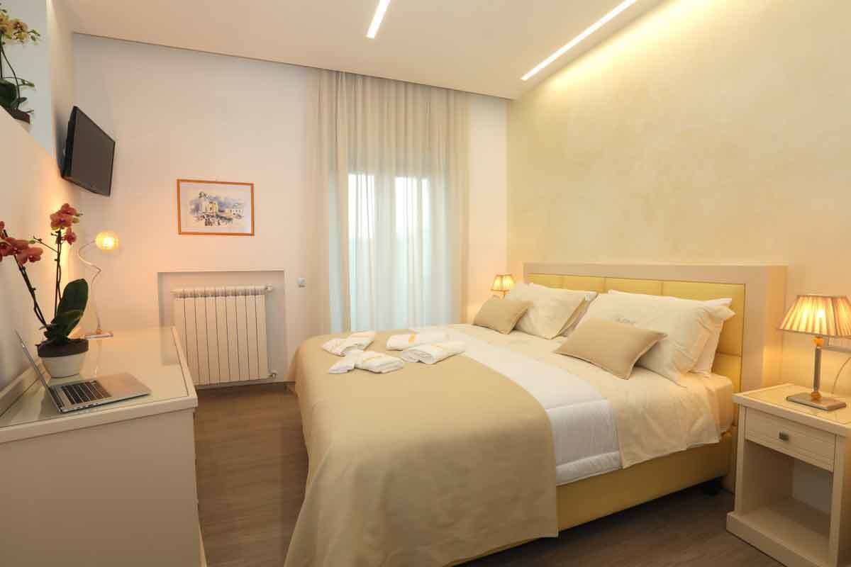 A hotel room with a bed, bedside tables, a desk with a laptop, a radiator, a TV mounted on the wall, and a window with curtains.