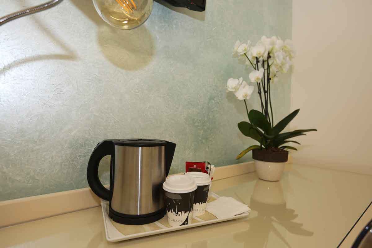 A coffee station with a stainless steel coffee pot, disposable cups, and a potted orchid on a countertop.