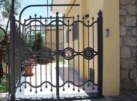 Una reja de hierro forjado con detalles decorativos, que da acceso a un edificio con un muro de piedra y una planta en maceta en primer plano.