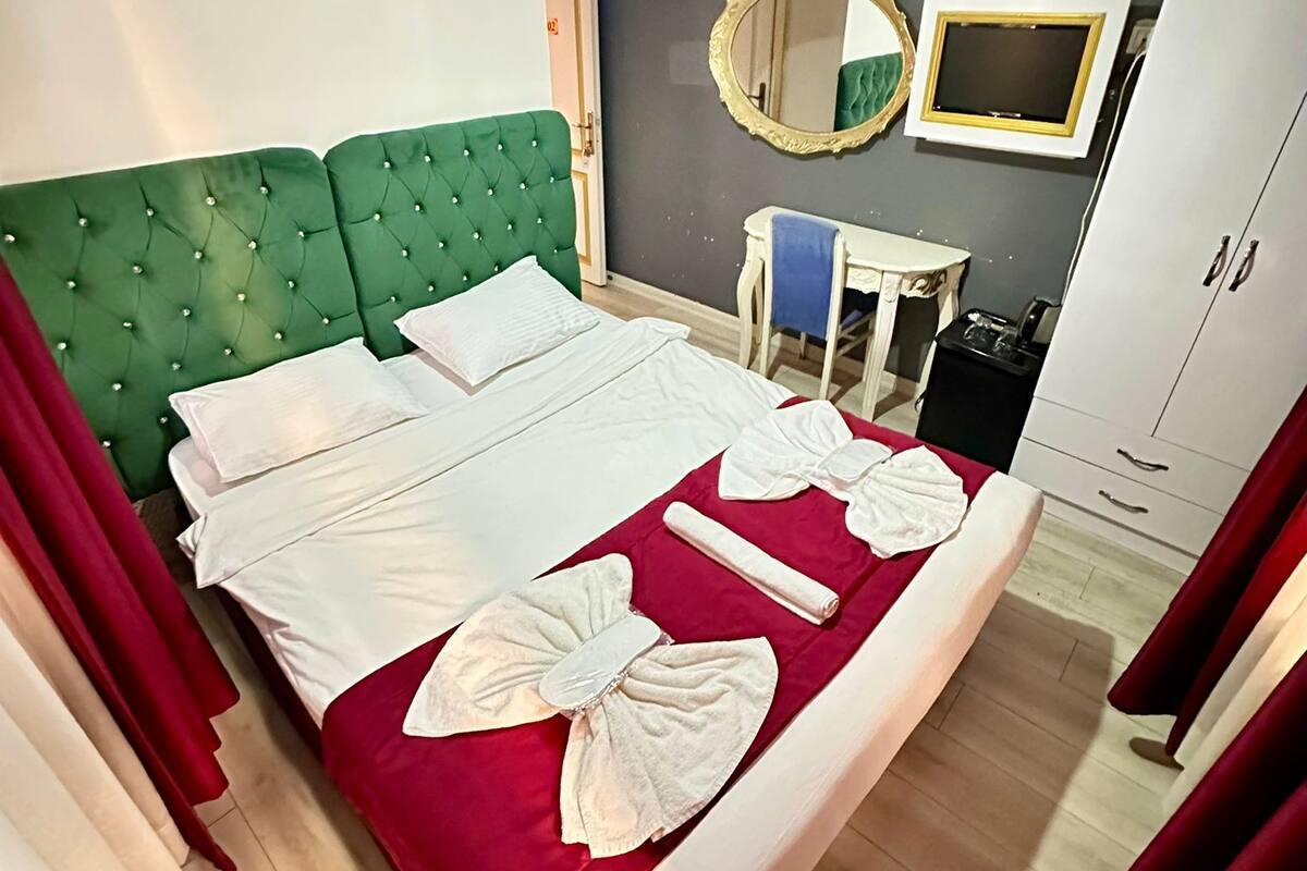 A hotel room with a green upholstered headboard, a bed with white linens, a red bedspread, and towels shaped like wings. There is a small table with a mirror and a white cabinet.