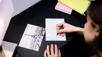 A person writing on a piece of paper at a table with a black surface, a yellow and pink notebook, and a black and white photo of a tower.