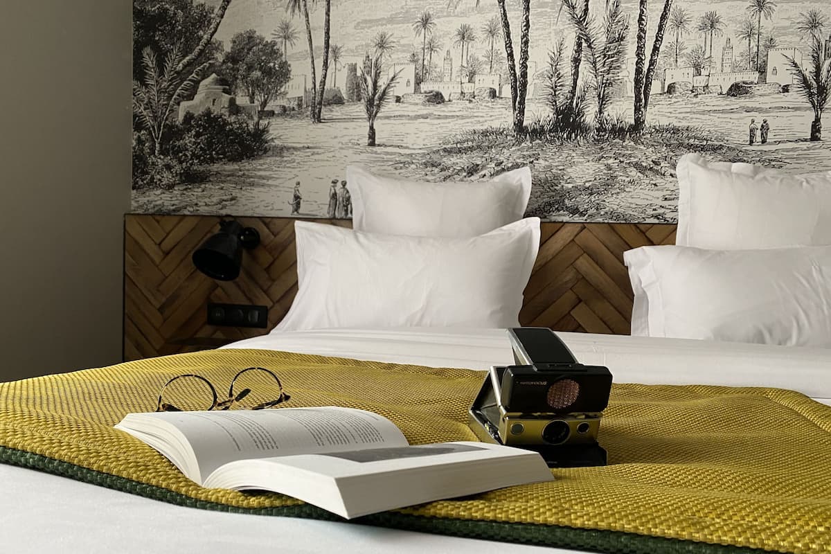 A hotel room with a bed, a large wall mural, a bedside table with a book and glasses, and a camera.
