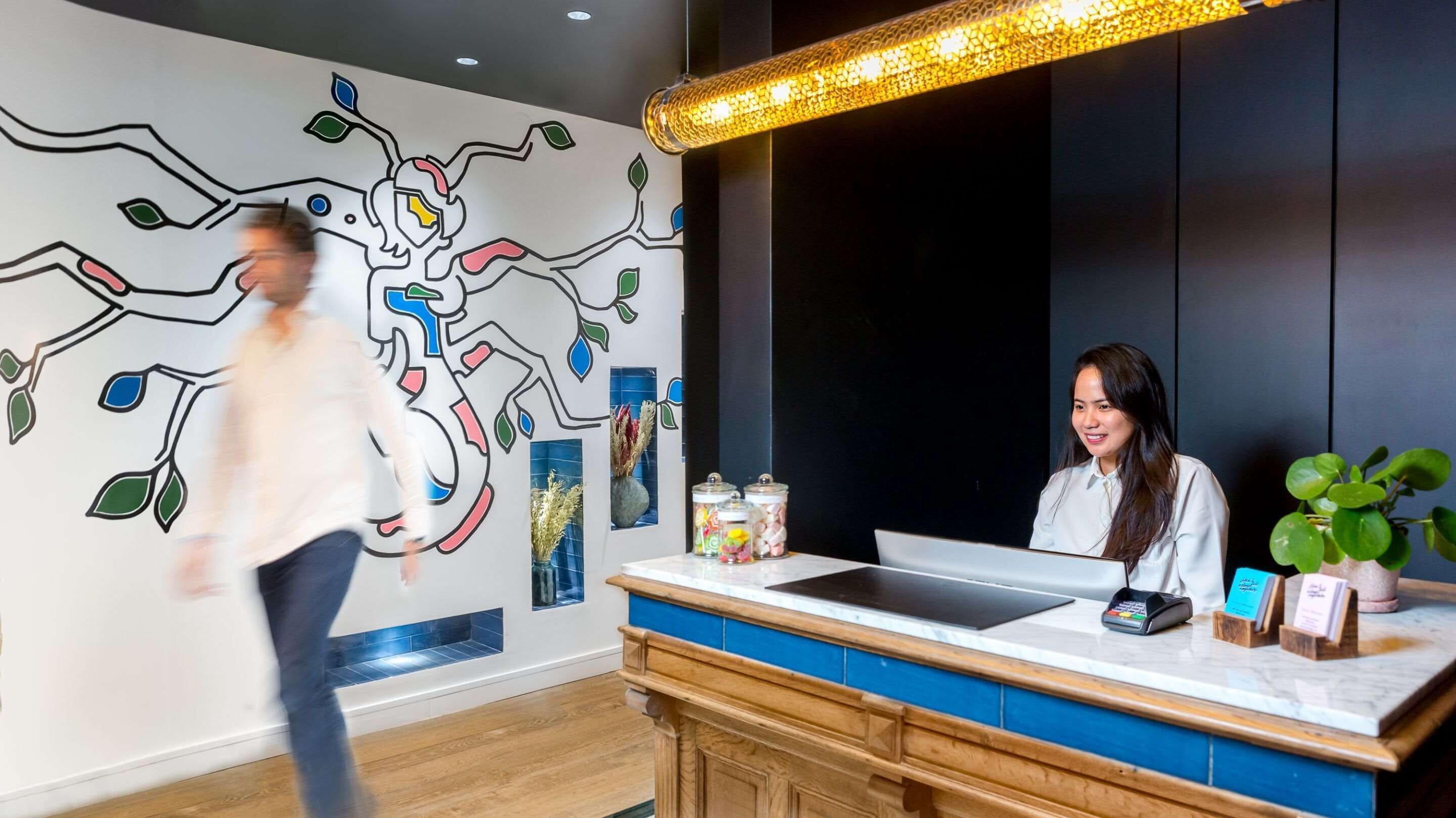 A reception area with a modern design, including a colorful wall mural, a reception desk with a computer, and a potted plant.