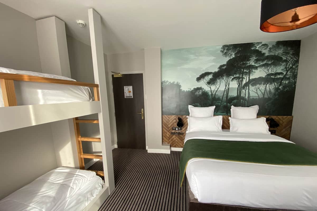 A hotel room with bunk beds, a large bed, a painting of a forest, and a wooden headboard.