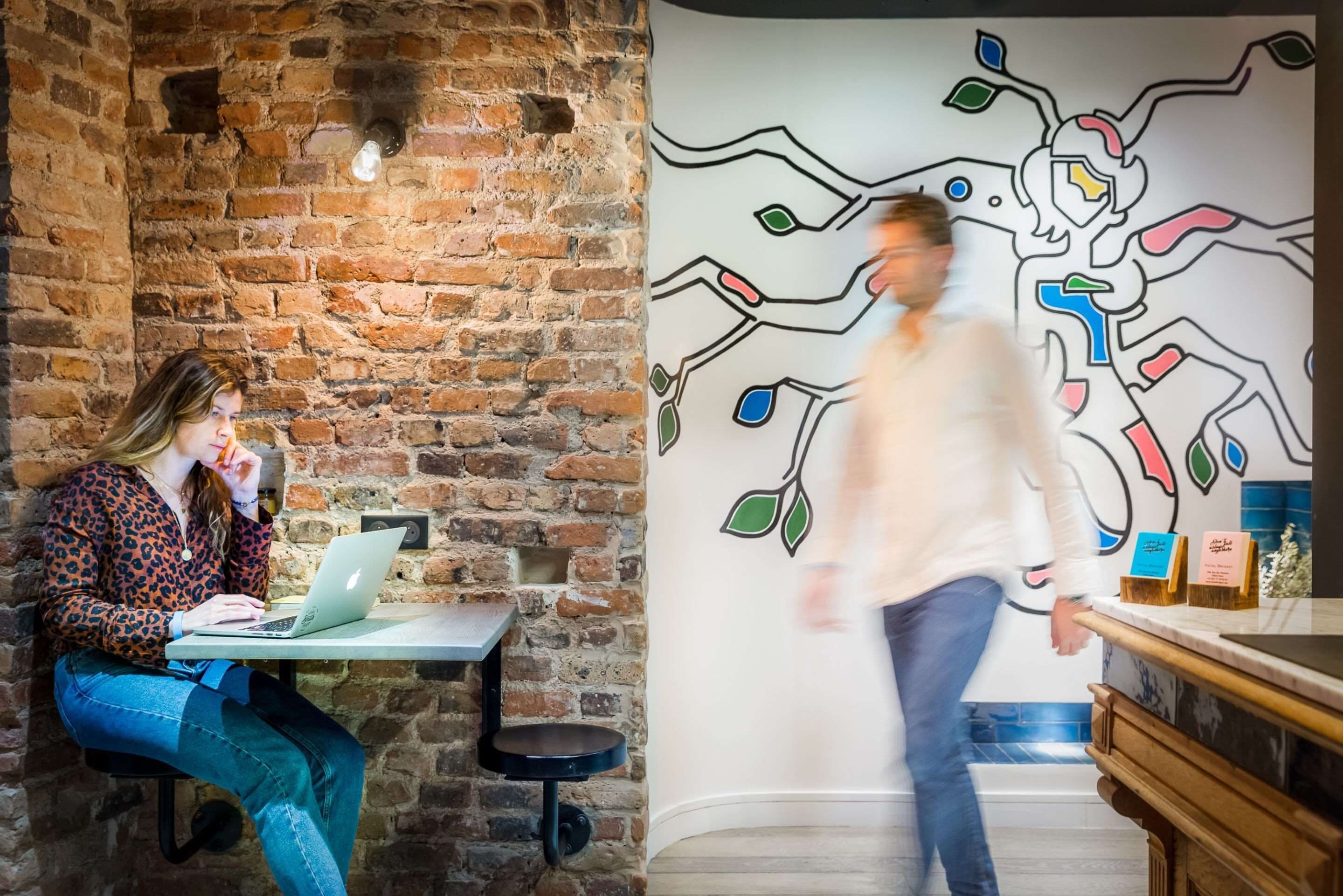 A person sitting at a table with a laptop in a room with a brick wall and a colorful abstract mural.