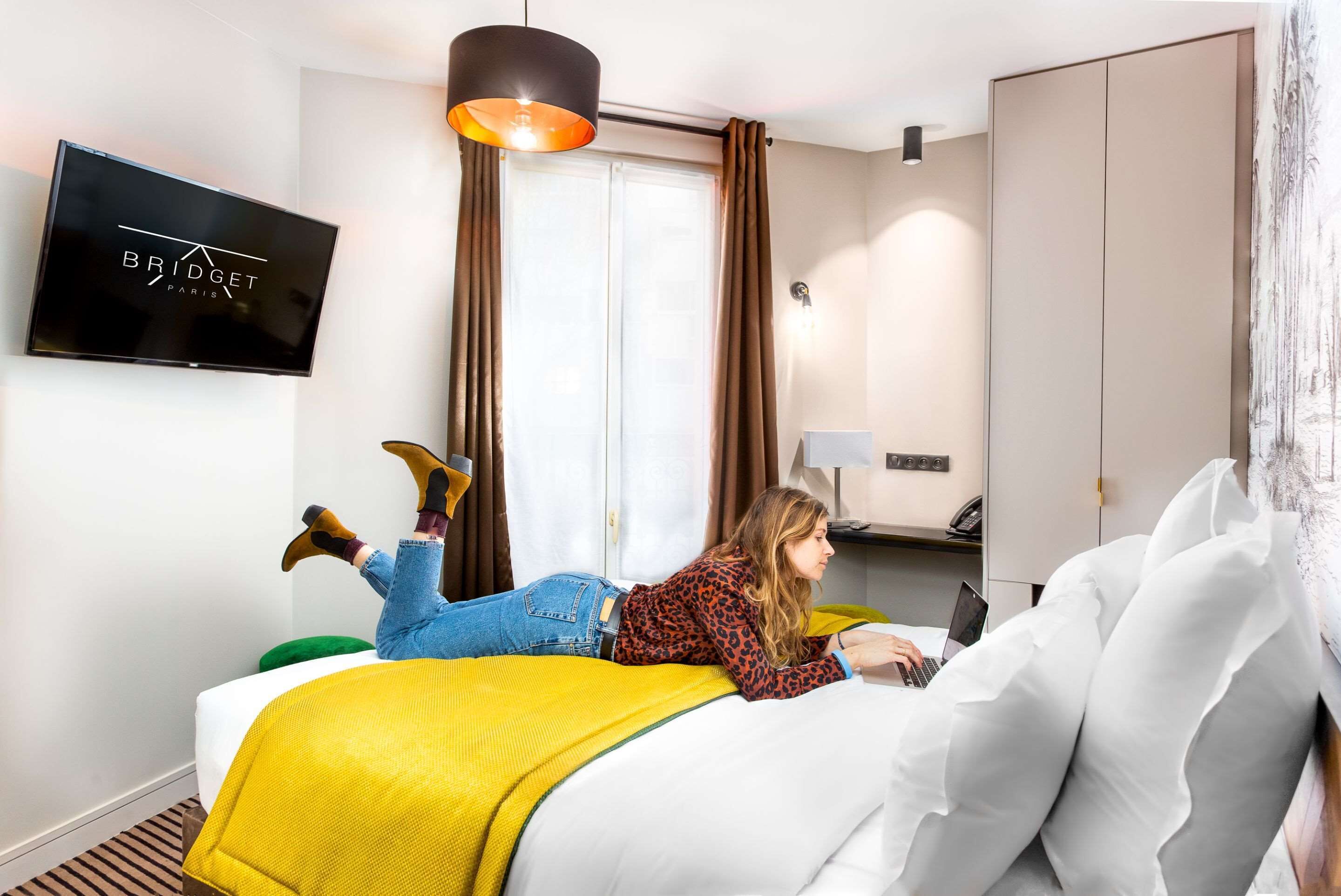 A hotel room with a bed, a TV mounted on the wall, a desk with a lamp, and a wardrobe.