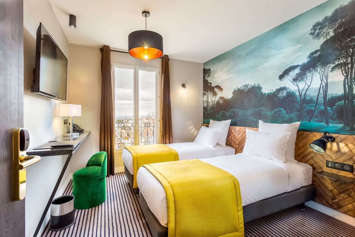 A hotel room with two beds, a desk, a TV, a painting, and a view of the outdoors.