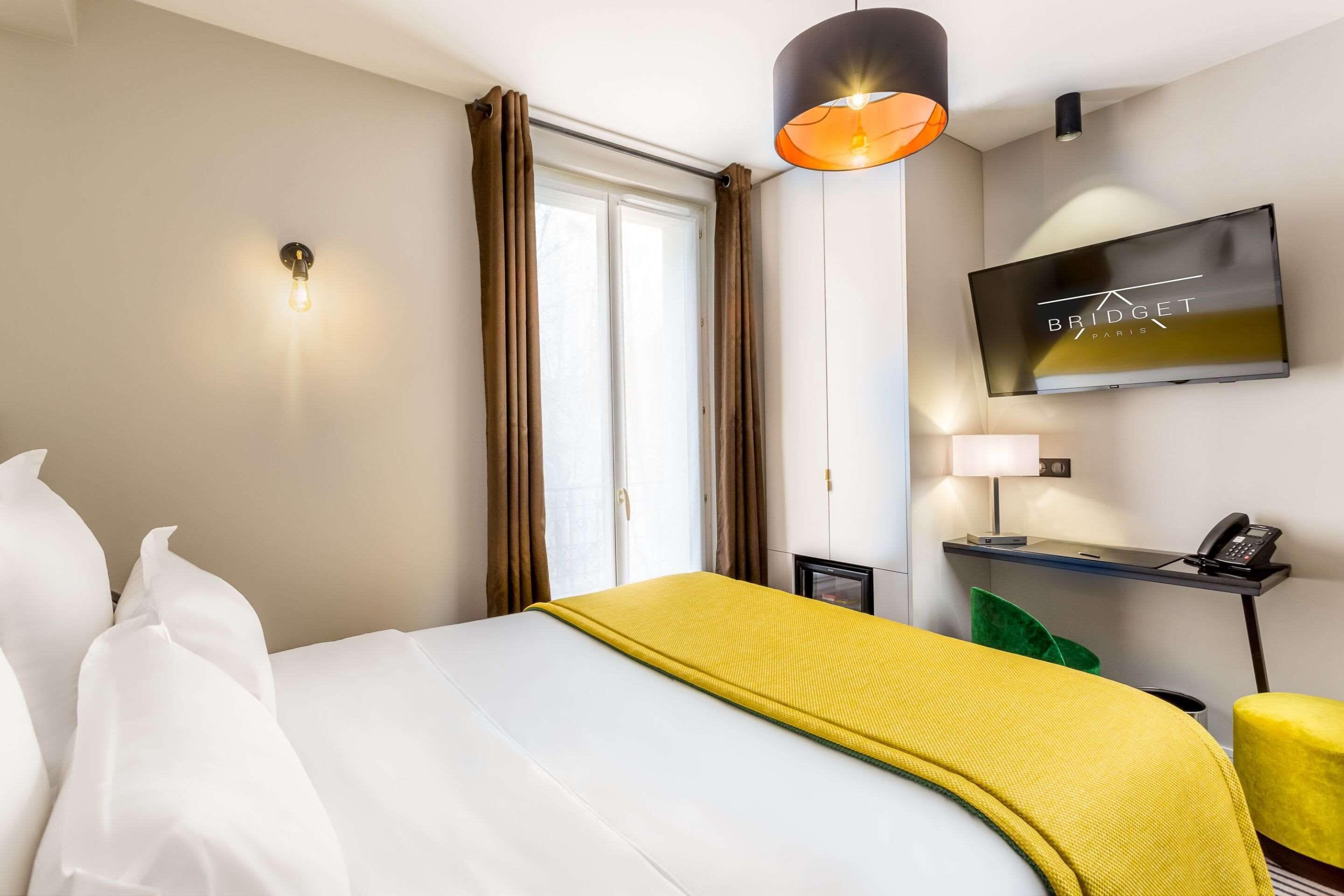 A hotel room with a bed, a TV mounted on the wall, a desk with a phone, and a window with curtains.