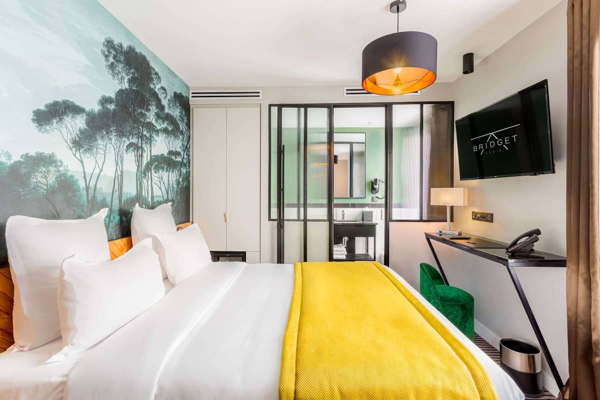 A hotel room with a bed, a desk, a TV, and a painting of trees.