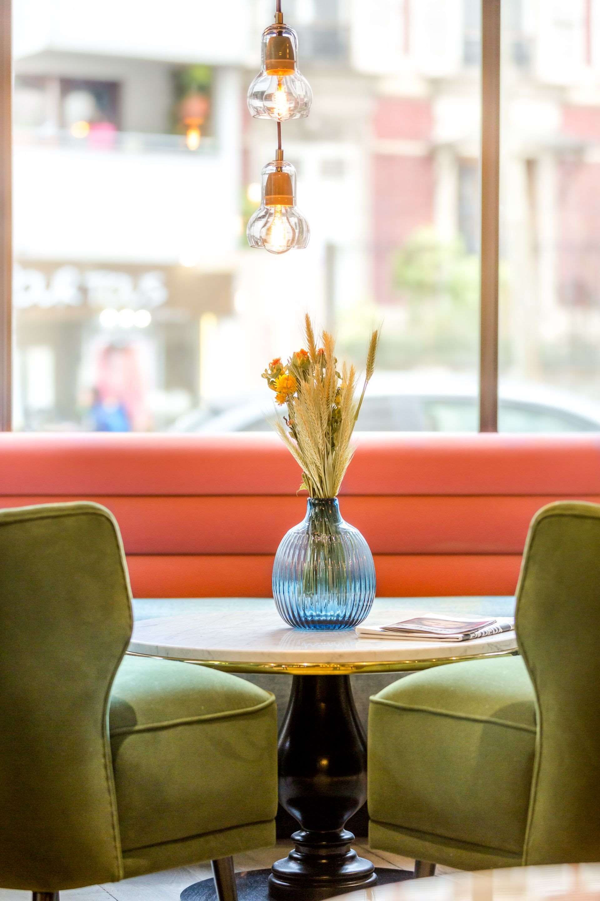 A table with a vase of flowers, green chairs, and hanging light fixtures in a brightly lit room with a red bench and a window view.