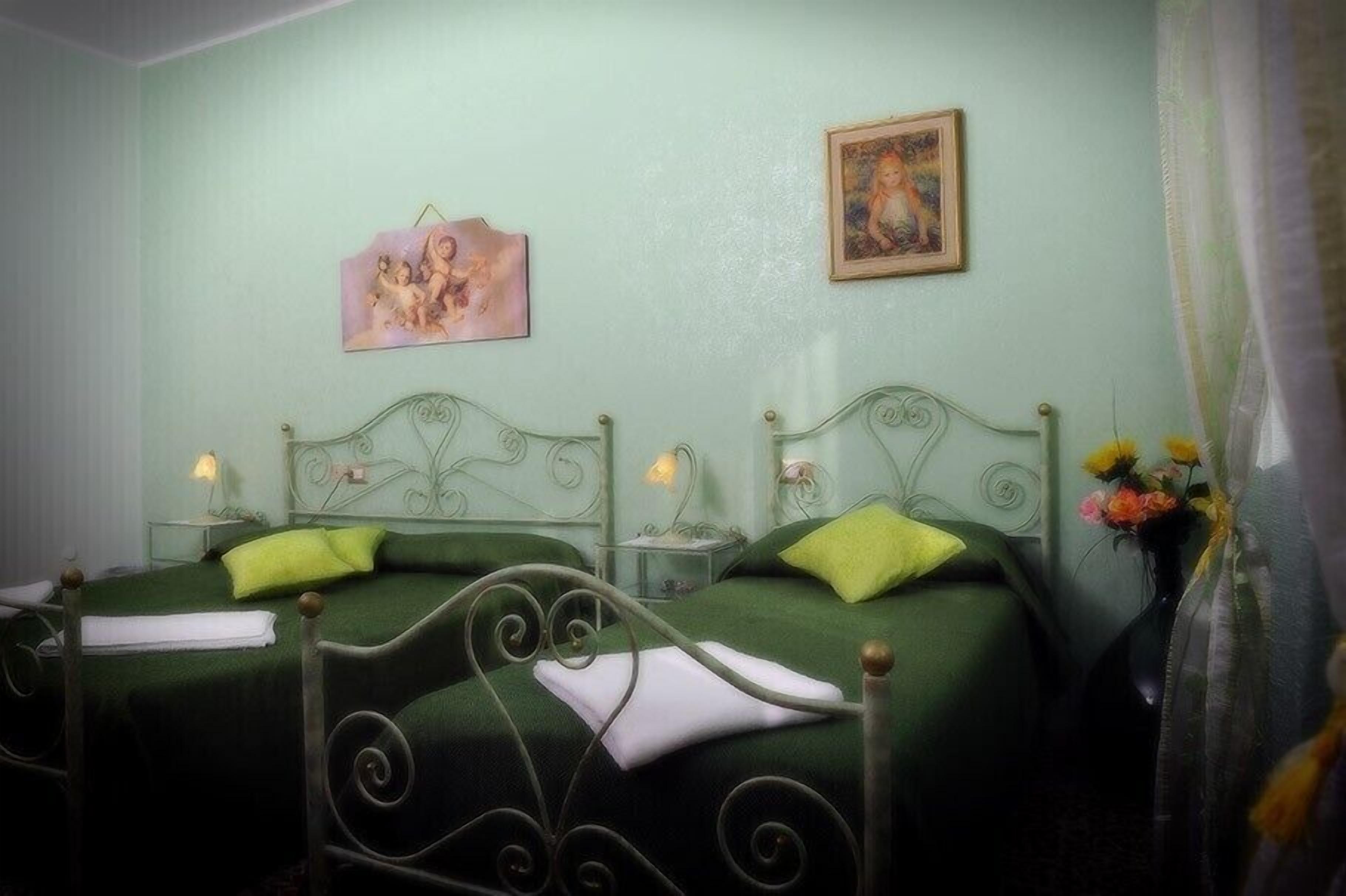 A room with two beds, each with green bedding and decorative pillows. There are bedside tables with lamps and a painting on the wall.