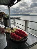 Una mesa con fruta y dispensador de agua en un balcón con vista al agua.