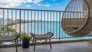 Un balcón con sillas de mimbre, una hamaca colgante y una planta en maceta, con vistas a la playa y al paisaje urbano.