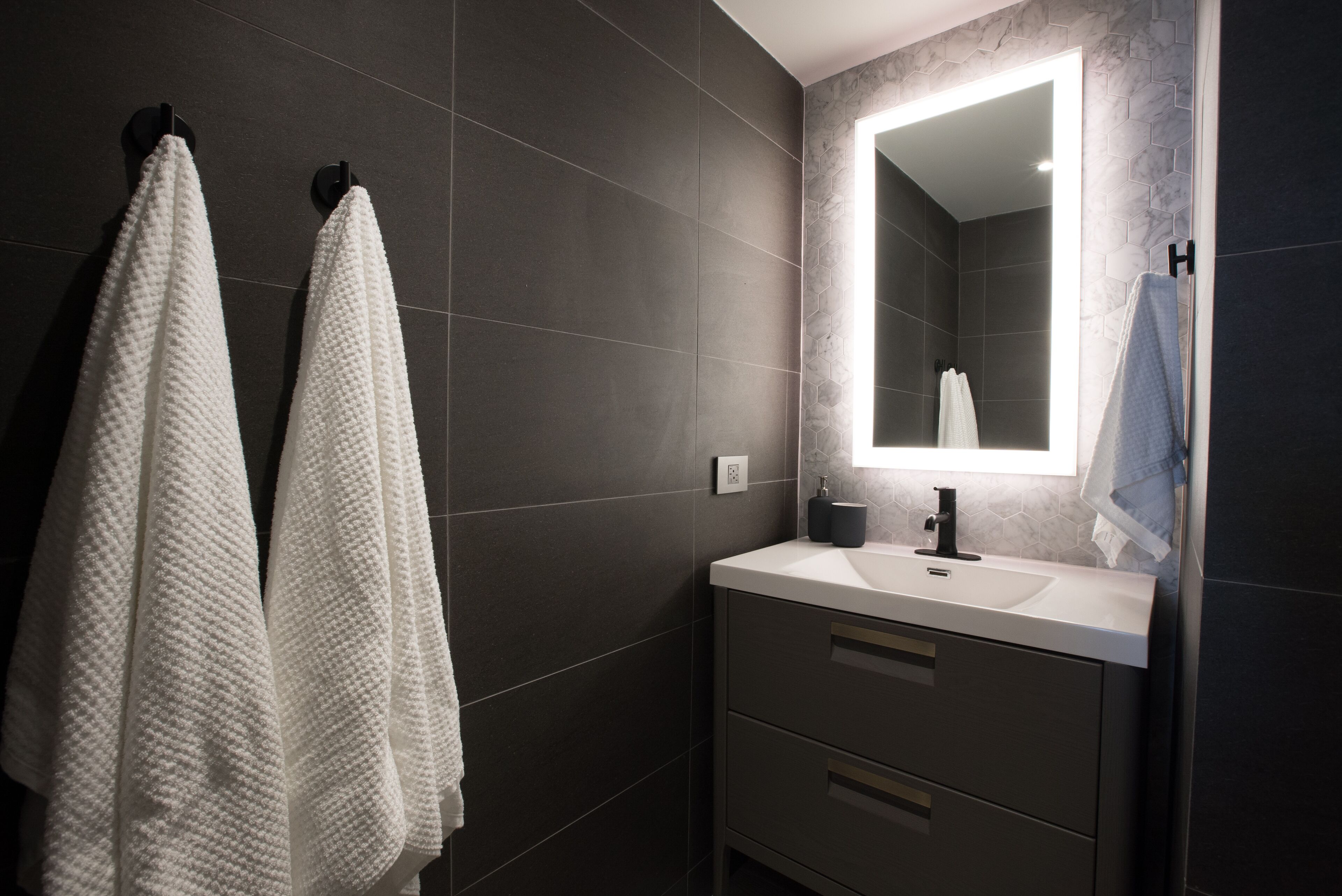Une salle de bain moderne avec des carreaux de couleur sombre, un lavabo blanc et deux serviettes suspendues à des crochets.