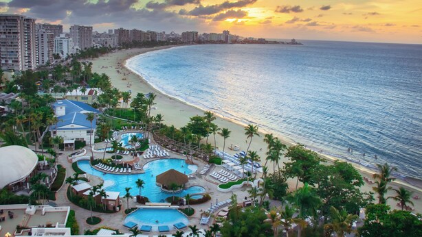 Un resort con una amplia piscina, acceso a la playa y edificios altos al fondo.