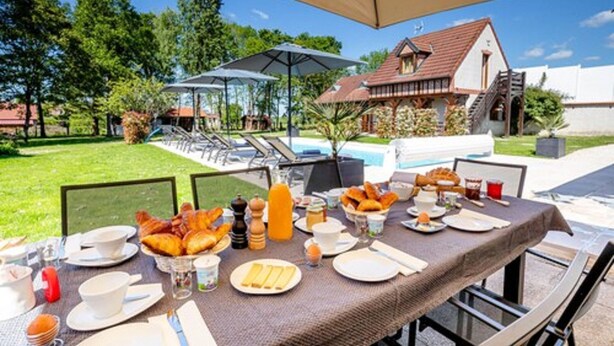 An outdoor dining area with a table set for breakfast, surrounded by umbrellas and chairs, with a pool and a building in the background. - L'Orée du Bois Bourgogne - Adults Only (Saint-Nicolas-lès-Cîteaux)