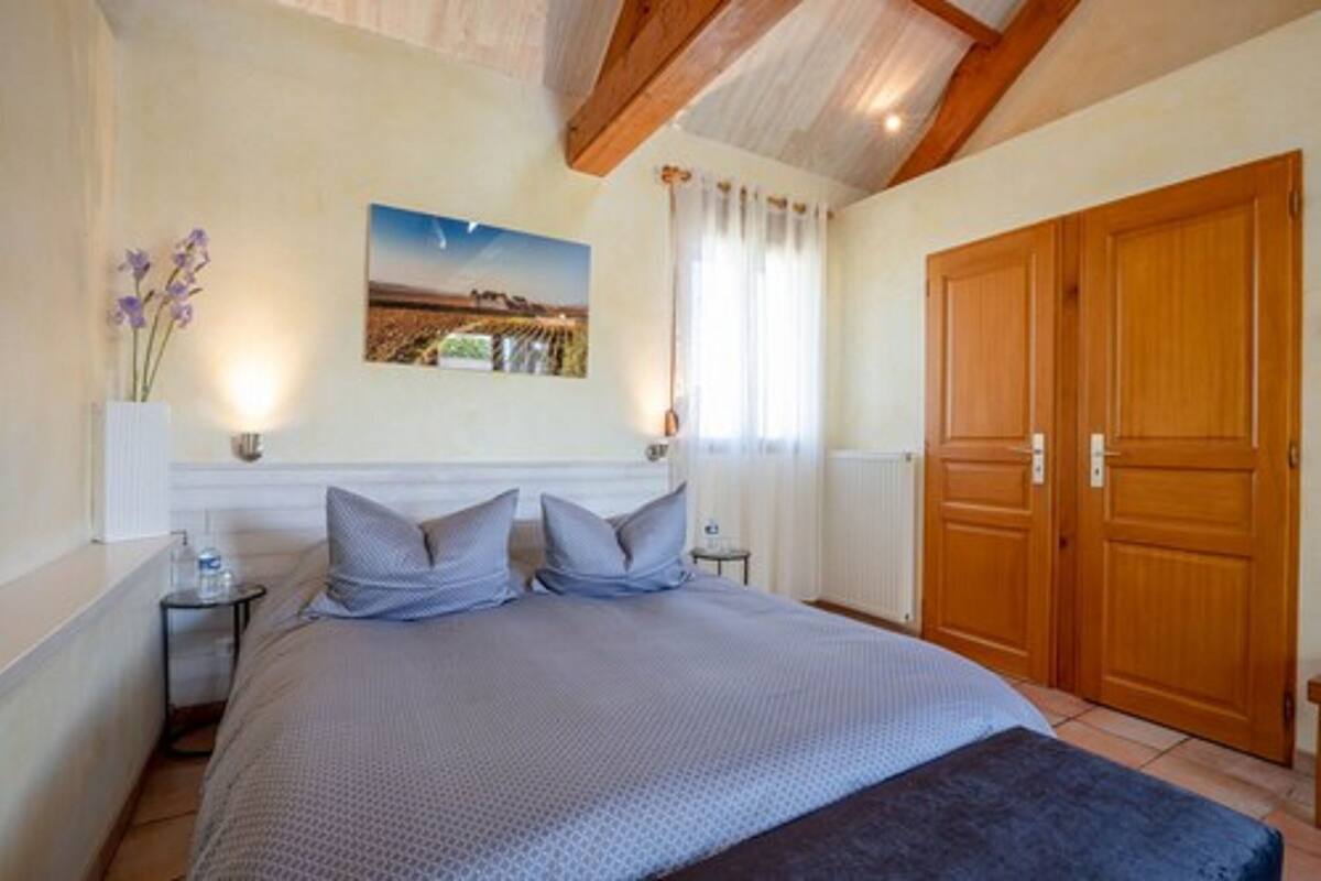 Une chambre à coucher comprenant un lit, une table de chevet, un tableau accroché au mur, une fenêtre avec des rideaux et une porte en bois.