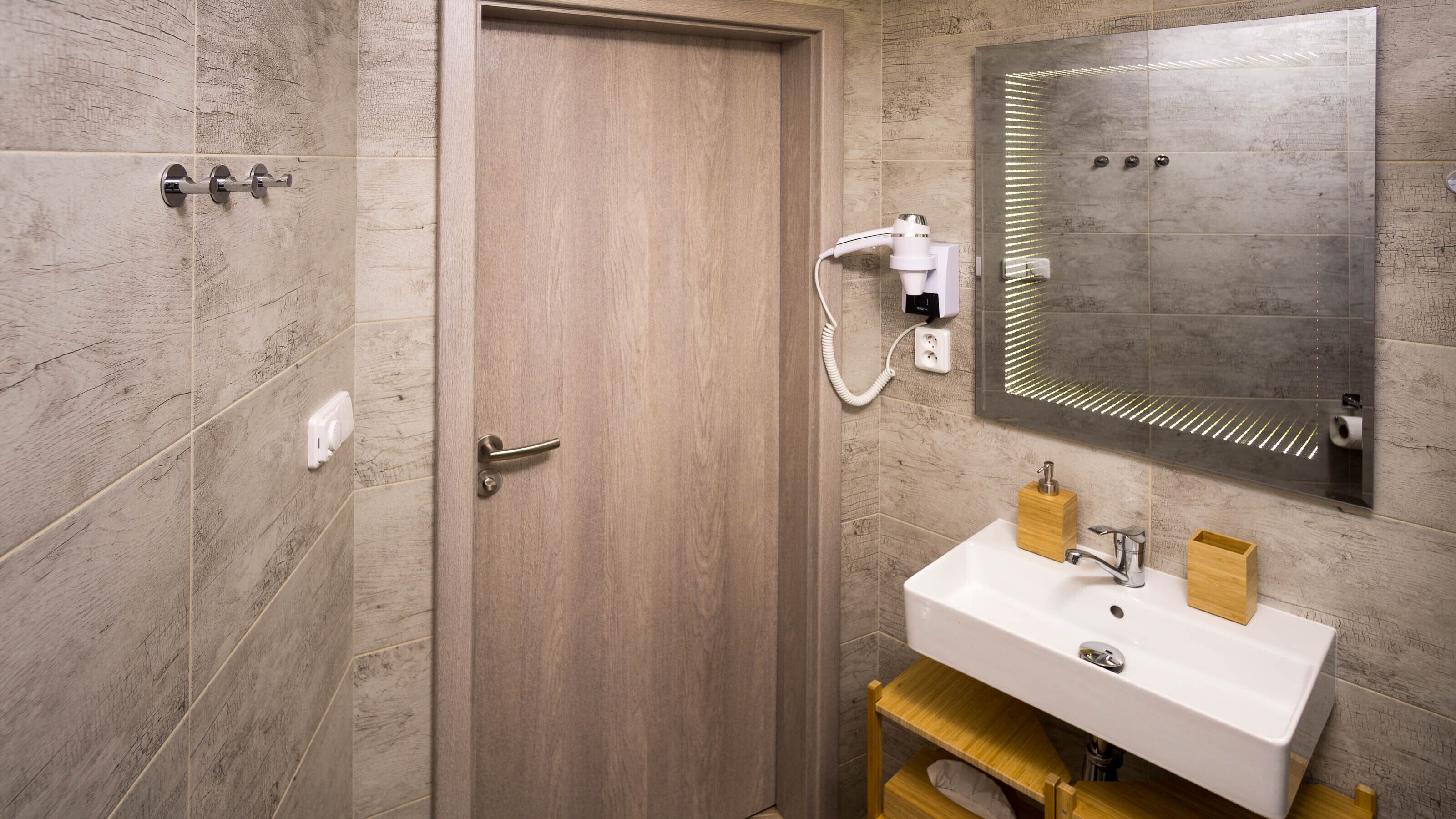 A bathroom with a wooden door, a white sink with a wooden soap dispenser, a mirror with LED lights, and a towel rack.