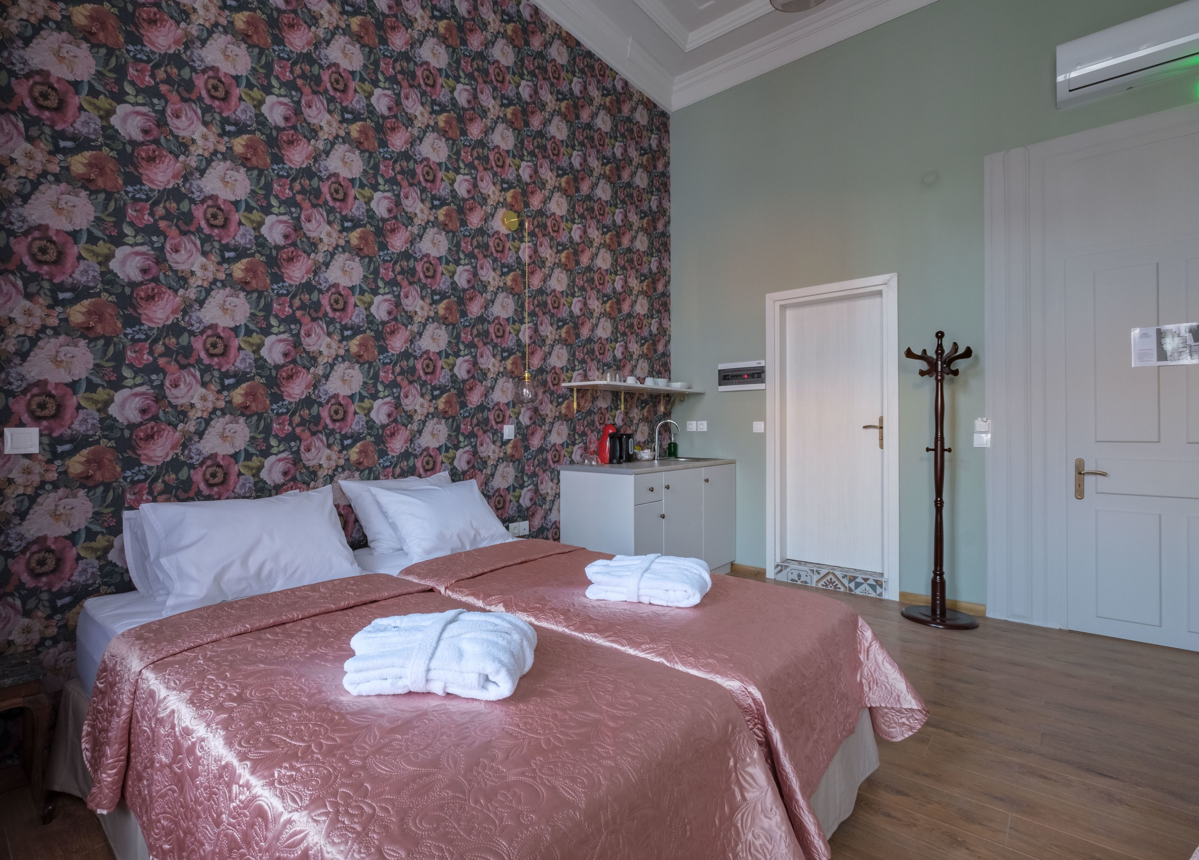 Une chambre avec un papier peint à motifs floraux, un lit recouvert d’une couverture rose, deux serviettes blanches, une table de chevet avec une bouilloire et un porte-manteau.