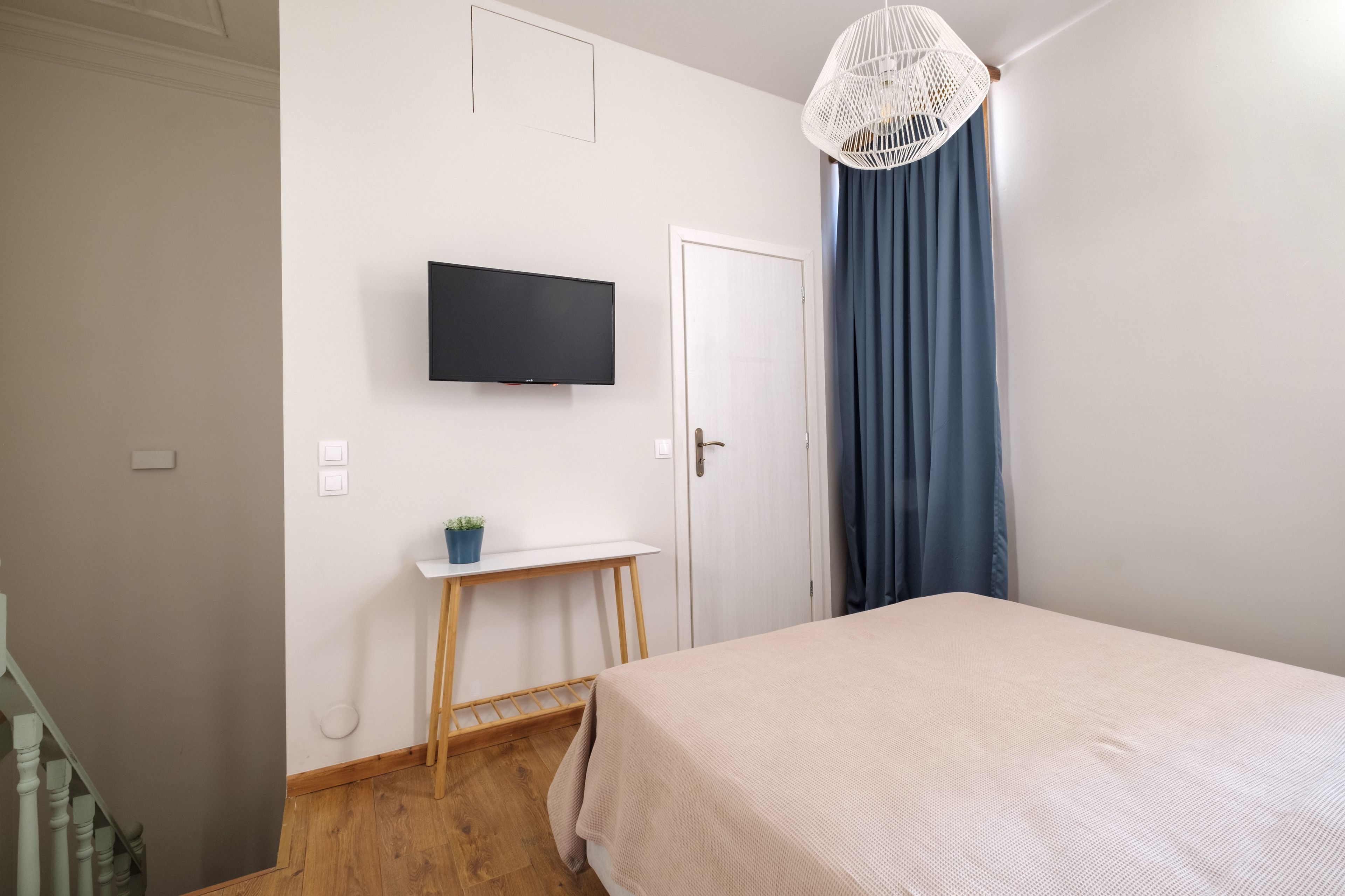 A bedroom with a bed, a TV mounted on the wall, a small table with a plant, and a door with curtains.