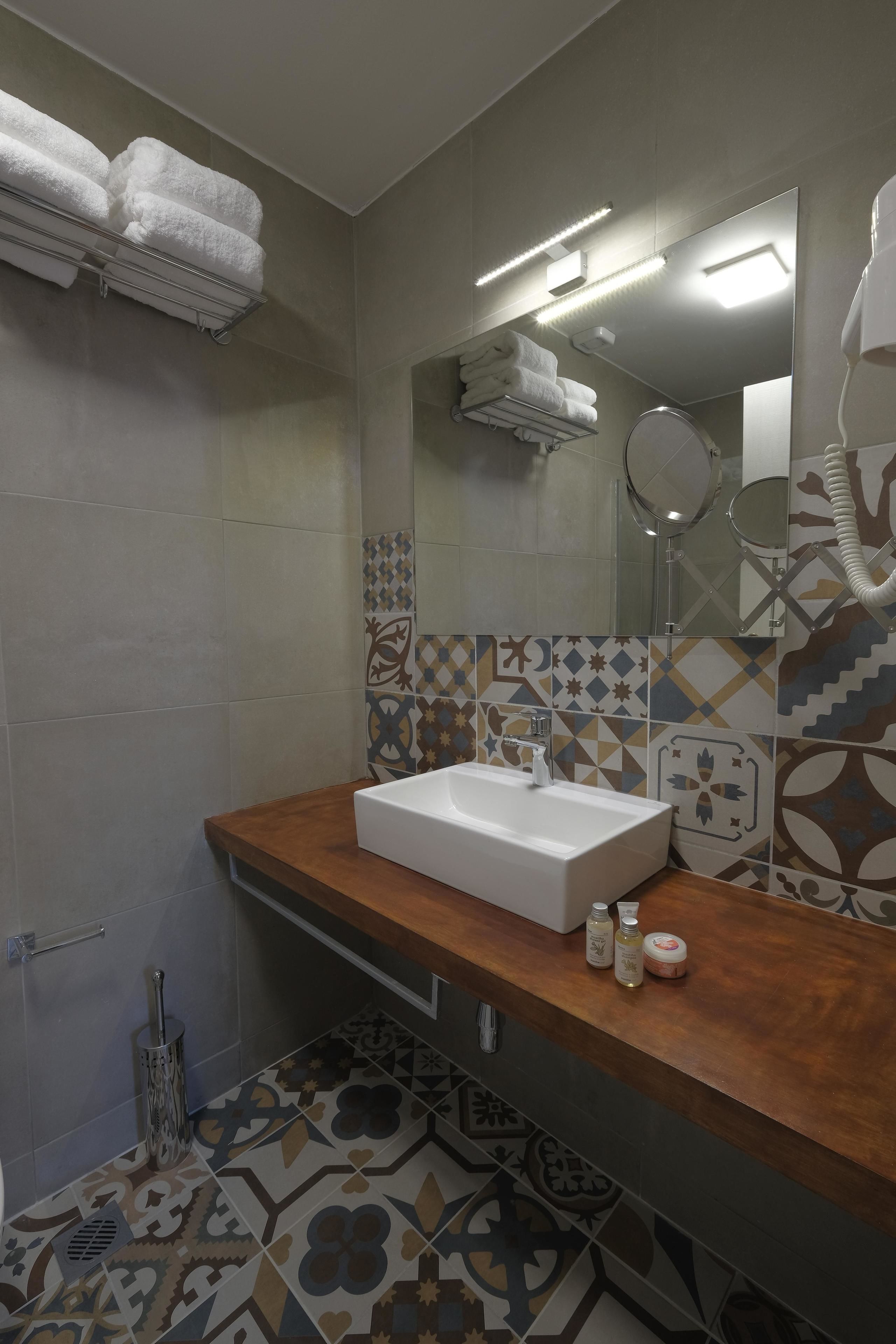 A bathroom with a wooden vanity, a white sink, and a mirror. There are towels on a shelf and a shower area visible in the background.