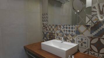 A bathroom with a wooden vanity, a white sink, and a mirror. There are towels on a shelf and a shower area visible in the background.