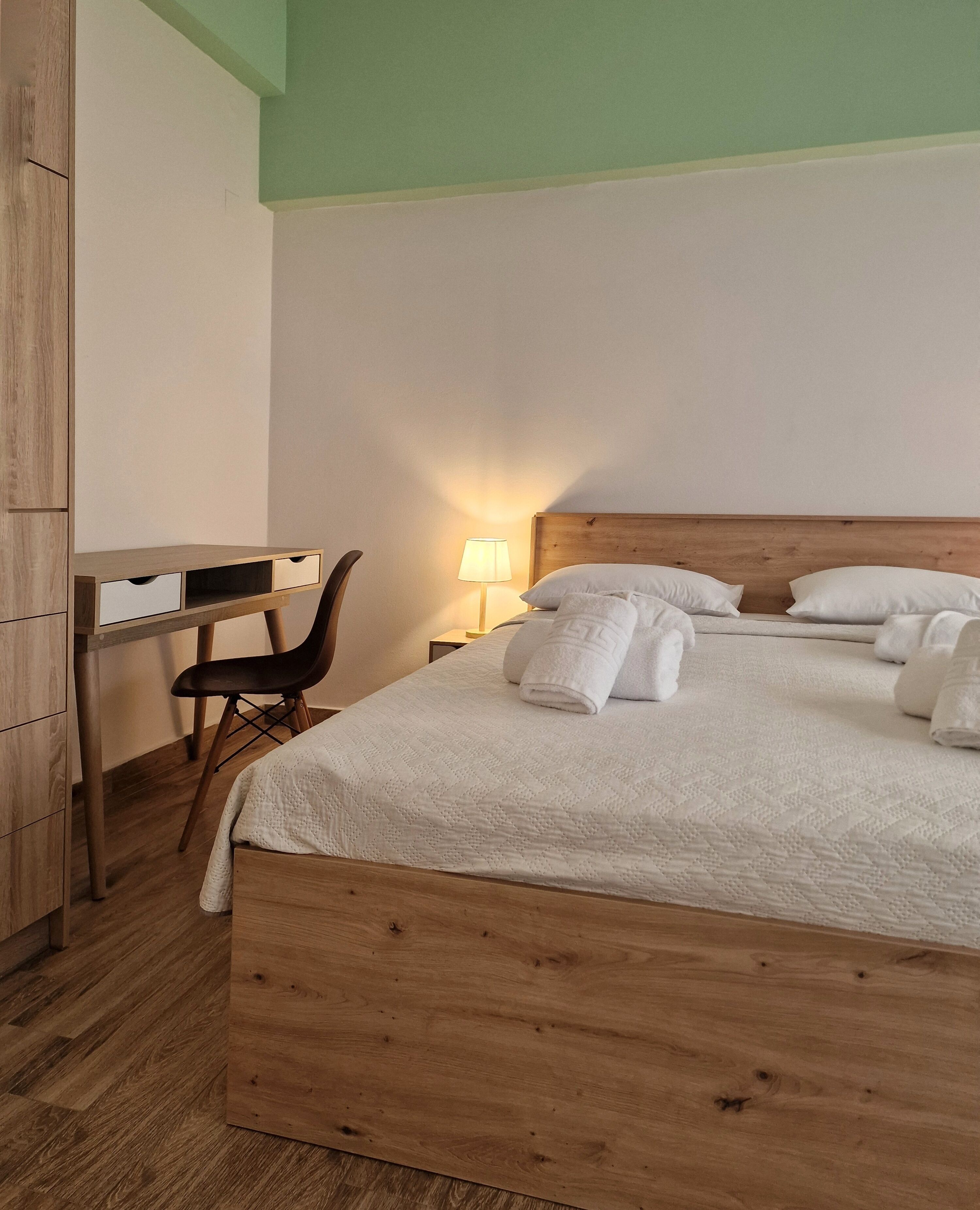 Una cama bien hecha con sábanas y almohadas blancas, un escritorio de madera con cajones, una silla y una lámpara de noche.