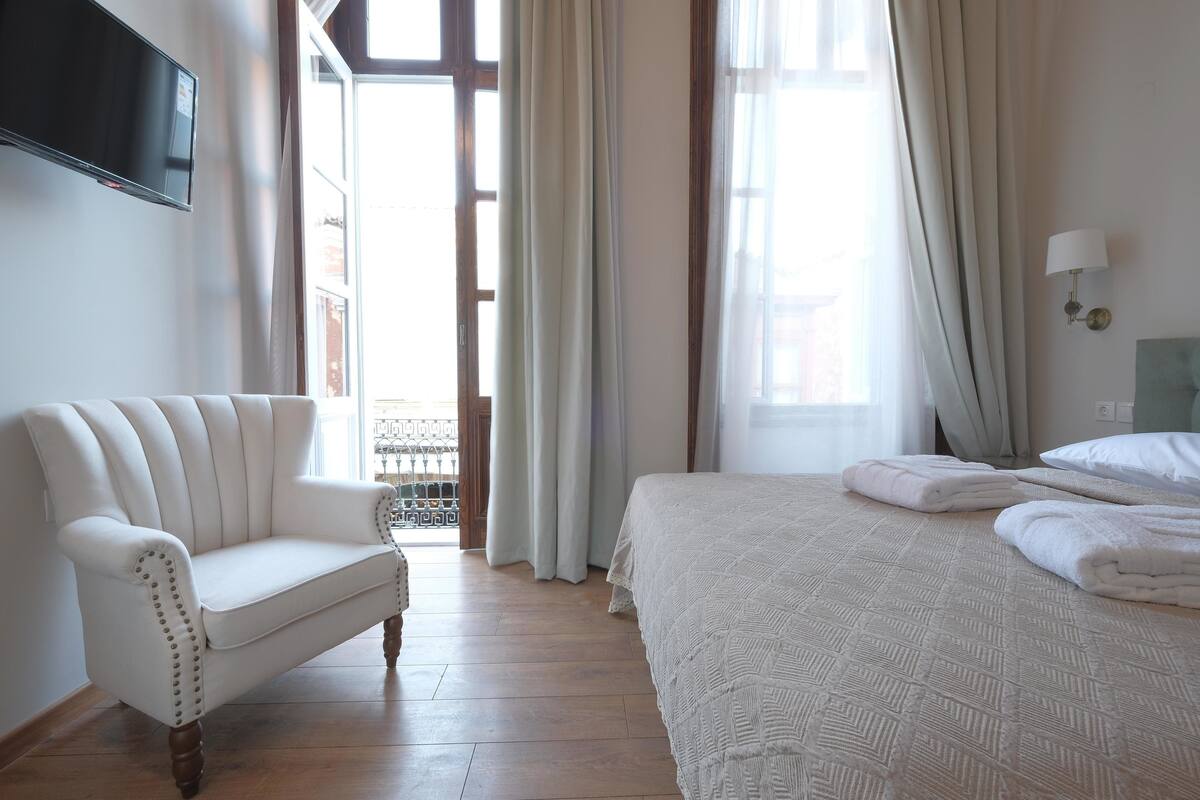 A hotel room with a white sofa, a neatly made bed with folded towels, a flat-screen TV mounted on the wall, and a window with curtains.