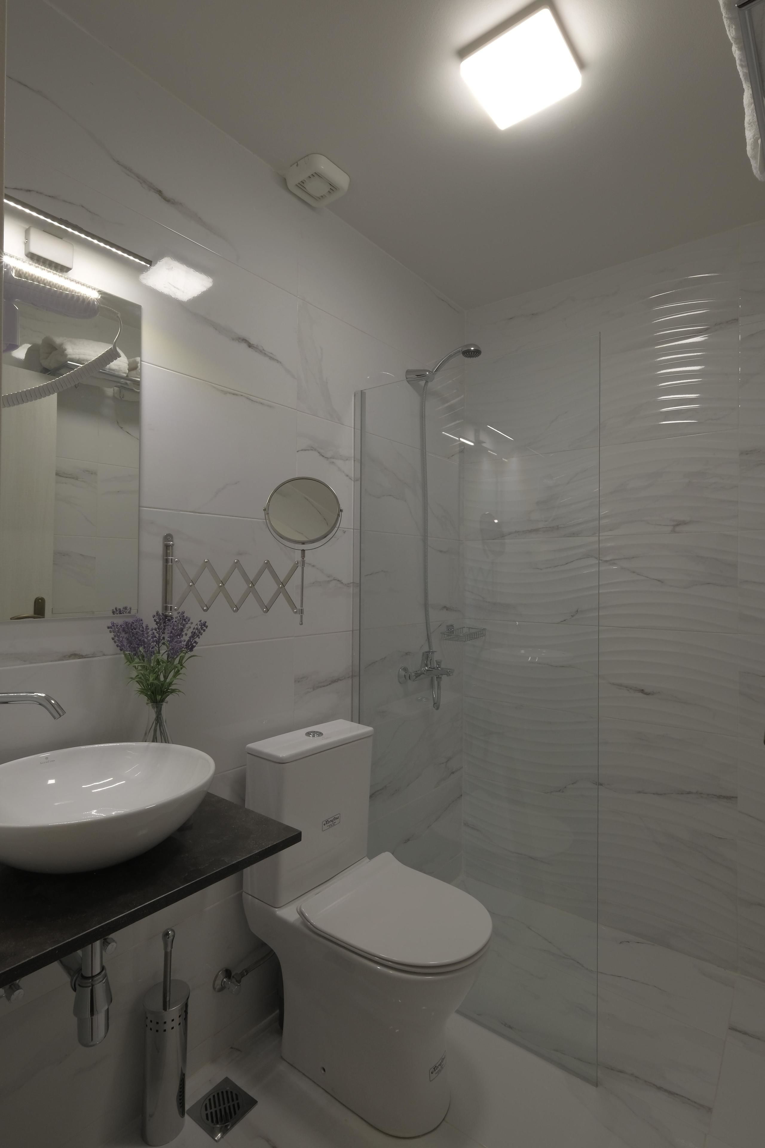 Un baño moderno con lavamanos, espejo y retrete blancos, paredes de mármol y zona de ducha.