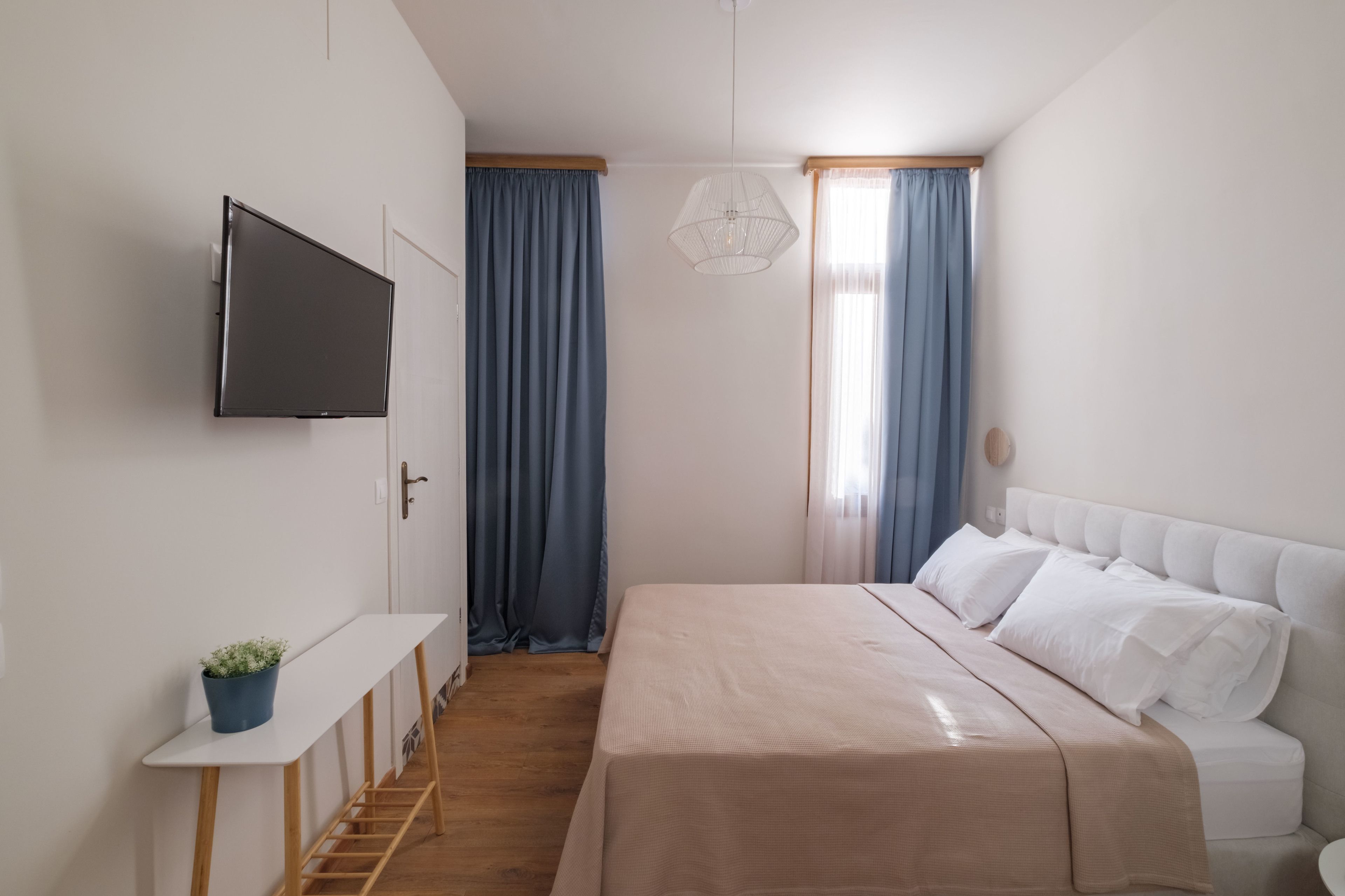 Un dormitori modern amb un llit, un televisor fixat a la paret, una taula petita amb una planta i una finestra amb cortines.