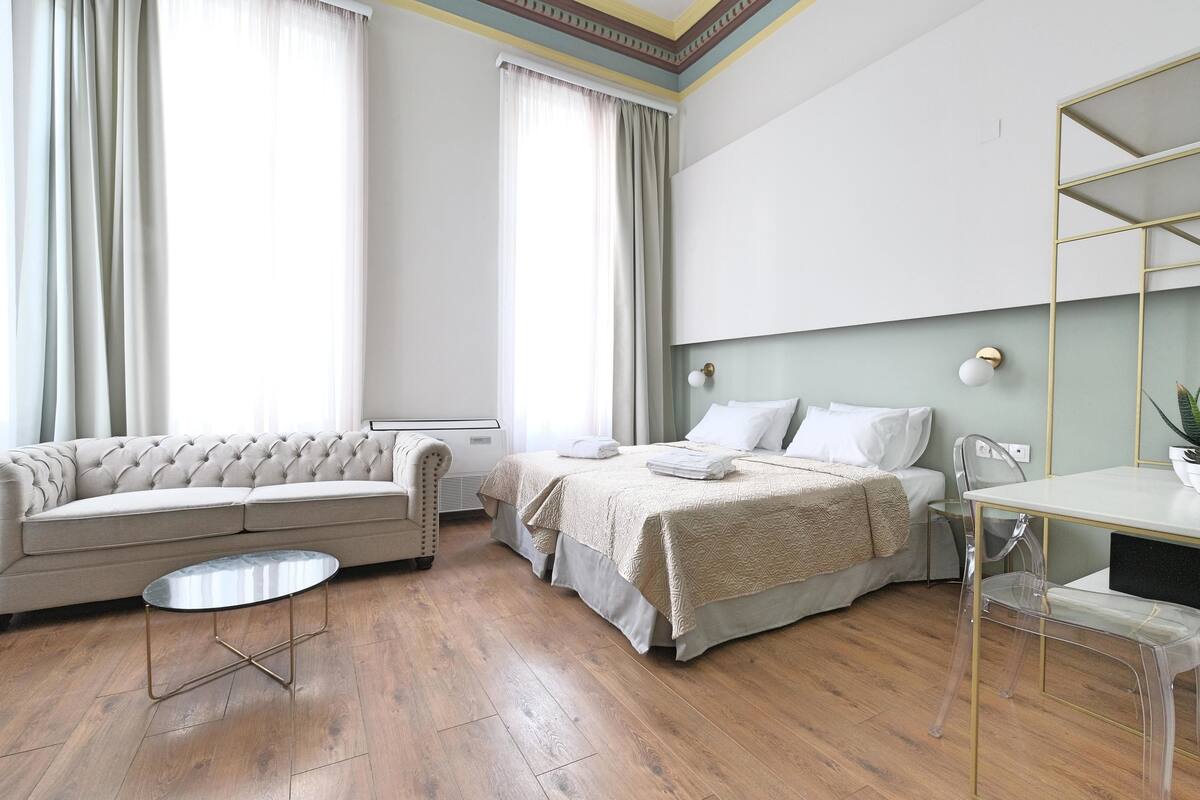 A modern hotel room with a sofa, a bed with a beige bedspread, a clear glass table, and a wooden floor.