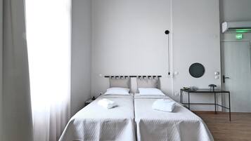 A hotel room with two beds, a bedside table, a round mirror, and a door with an emergency exit sign.