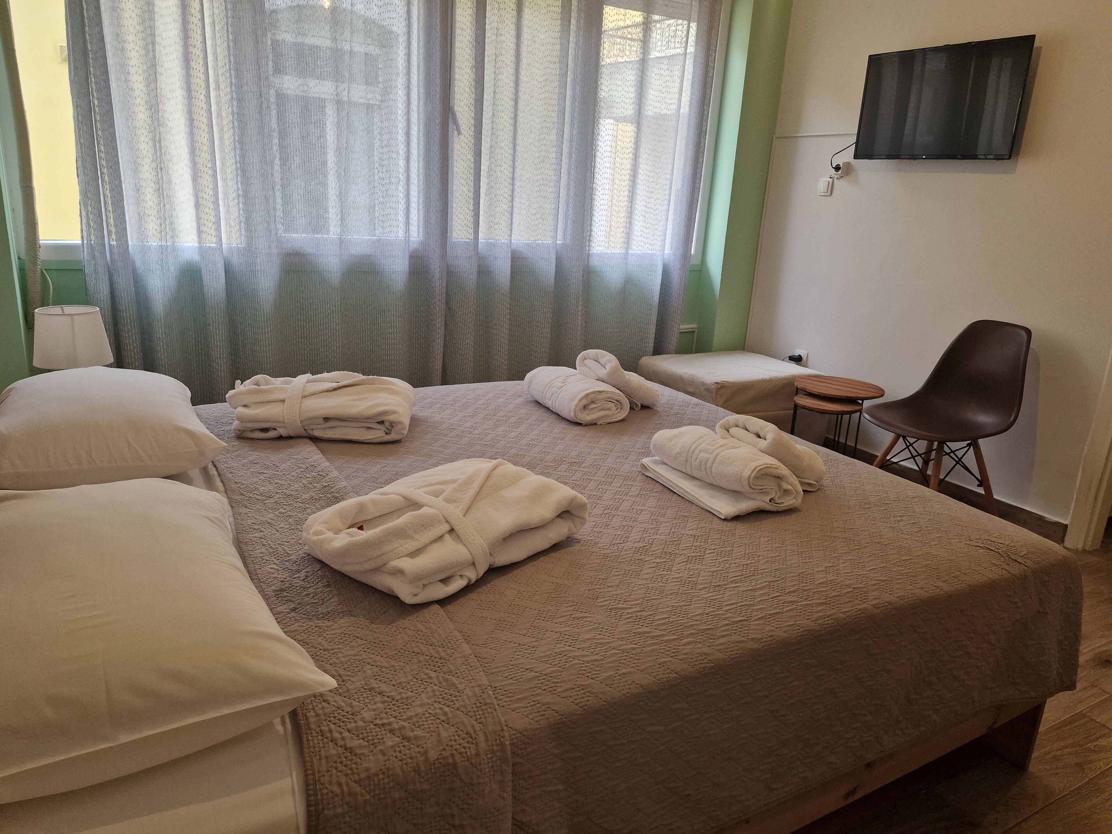 Une chambre d’hôtel avec un grand lit, deux tables de chevet, une chaise, un téléviseur fixé au mur et une fenêtre avec des rideaux légers.