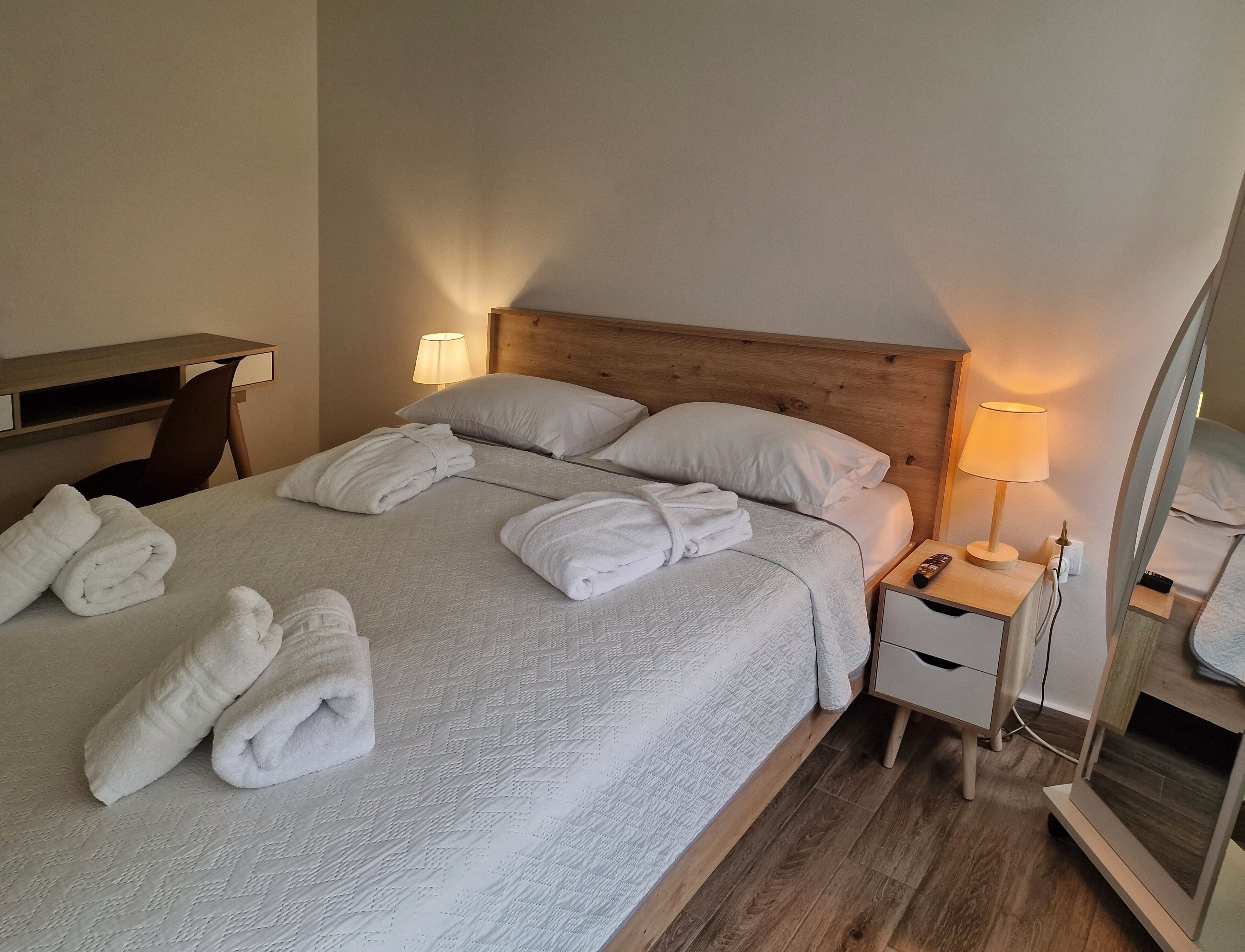 Una cama bien hecha con sábanas y toallas blancas, un cabecero de madera, lámparas de noche y una mesita auxiliar con un control remoto.