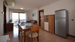 A compact kitchen with a refrigerator, wooden cabinets, and a dining area with chairs and a table. - Residence Porta del Mare (Lignano Sabbiadoro)
