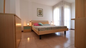 A bedroom with a bed, nightstands, a wooden wardrobe, a framed picture on the wall, and a window with curtains. - Residence Porta del Mare (Lignano Sabbiadoro)