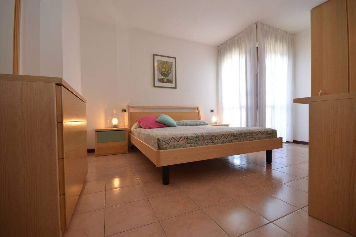 Ein Schlafzimmer mit einem Bett, Nachttischen, einem Holzschrank, einem gerahmten Bild an der Wand und einem Fenster mit Vorhängen.
