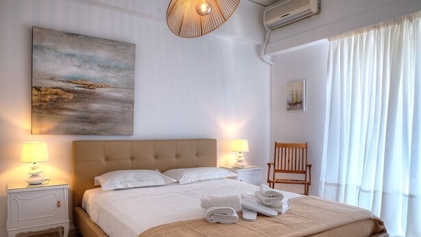A neatly made bed with white linens, a beige headboard, and a bedside table with a lamp. - L' apartment blanc dans le centre de Glyfada (Glyfada)
