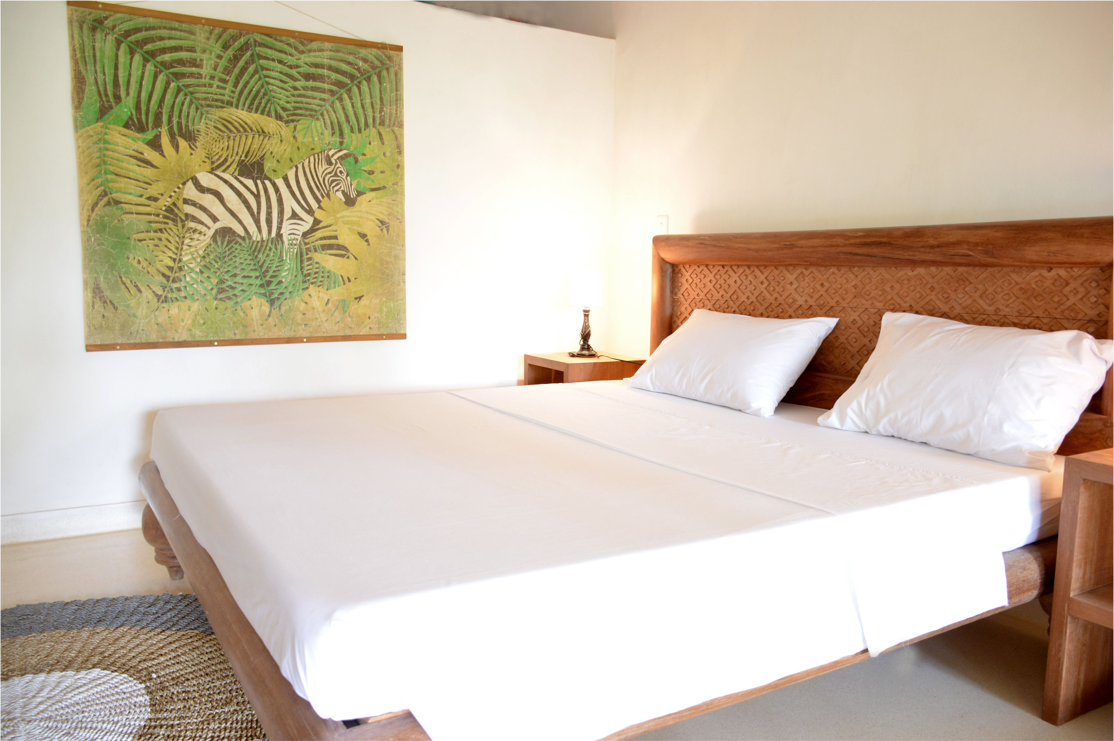 Un dormitorio con una cama grande, cabecera de madera, ropa de cama blanca, una mesita de noche con lámpara y un cuadro de cebra en la pared.