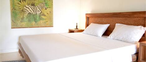 Un dormitorio con una cama grande, cabecera de madera, ropa de cama blanca, una mesita de noche con lámpara y un cuadro de cebra en la pared.